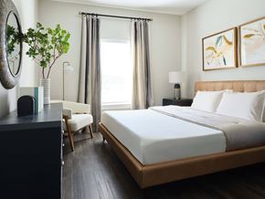 Panama City apartment for rent - Panama City, FL Pet-Friendly Luxury Apartments - The Kelly at 23rd - Bedroom with Vinyl Wood-Inspired Plank Flooring, Queen-Size Bed, Nightstand with Lamp, Dresser, Wall-Mounted Round Mirror, Armchair, Floor Lamp, and Large Window.