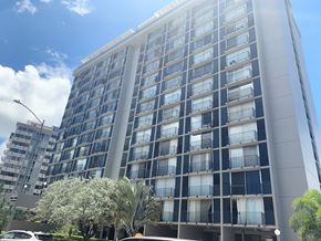 Honolulu apartment for rent - A tall building with many windows and balconies is surrounded by trees and a clear sky.