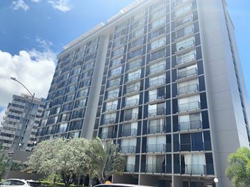 Honolulu apartment for rent - A tall building with many windows and balconies is surrounded by trees and a clear sky.