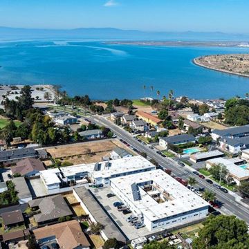 San Leandro apartment for rent - A large white building with a blue roof is surrounded by other buildings and palm trees.