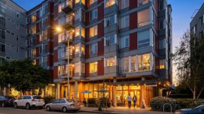 Seattle apartment for rent - A modern apartment building with cars parked in front and people walking on the sidewalk.