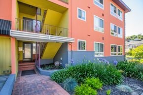 Oakland apartment for rent - A building with a yellow and pink exterior has a balcony and a small garden in front.