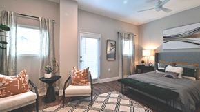 Mckinney apartment for rent - A residence bedroom at Bell Tower Flats. A large bed sits along the wall to the right, with end tables on either side. A door to the patio is to the left with a chair next to it.