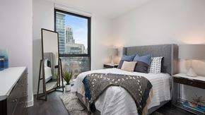Chicago apartment for rent - A bedroom in a Hubbard221 residence. The bed in on the right with end tables on both sides. There is a tall window past the bed with a standing mirror beside it. There is a dresser on the left wall.