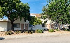 Grover Beach apartment for rent - A white house with a brown door and windows is surrounded by trees and bushes.