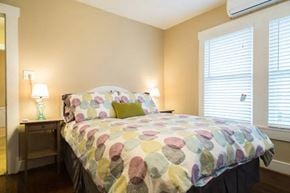 Fort Worth house for rent - A bed with a multicolored comforter and two green pillows sits in a room with a window and a nightstand with a lamp.