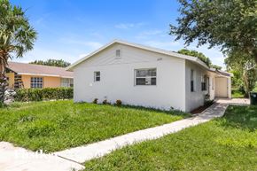 Greenacres house for rent - A white house with a brown roof is surrounded by green grass and trees.