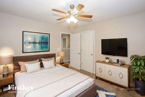 Charlotte house for rent - A bedroom with a bed, a ceiling fan, a nightstand with a lamp, a painting on the wall, a TV on a cabinet, and a potted plant.