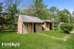 Arnold house for rent - A small house with a brown roof and a white fence is surrounded by green grass and trees.