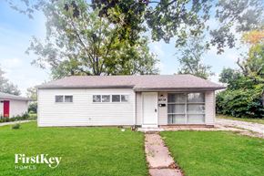 Indianapolis house for rent - A small white house with a red door is surrounded by green grass and trees.