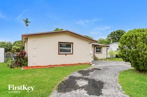 Sunrise house for rent - A beige house with a brown roof and a white fence is surrounded by greenery.