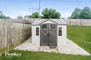 Greenwood house for rent - A small white shed with a grey door is situated in a grassy area with a wooden fence in the background.