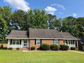Wilson apartment for rent - A house with a grey roof and a white door is surrounded by green bushes and trees.