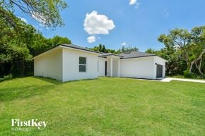 Deltona house for rent - A white house with a black roof is surrounded by green grass and trees.