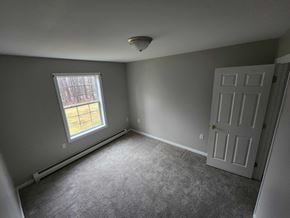 Gray apartment for rent - A room with a carpeted floor, a white door, and a window with a view of a building outside.