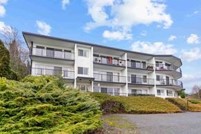 Nanaimo apartment for rent - A white building with balconies and windows is surrounded by greenery.