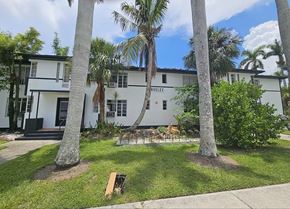 Fort Myers apartment for rent - A white building with a black door and windows is surrounded by palm trees.