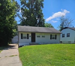 St. Louis house for rent - A small house with a grey roof and white siding is surrounded by a green lawn and trees.