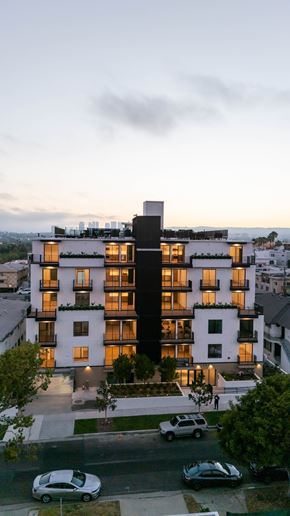 Los Angeles apartment for rent - A modern multi-story residential building with balconies and large windows is illuminated from within, with cars parked in front and a cloudy sky above.
