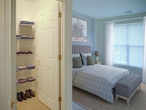 Silver Spring apartment for rent - A bedroom with a bed, pillows, a nightstand, a window with blinds, a rug, a door, a shelf with folded clothes, and a pair of shoes.