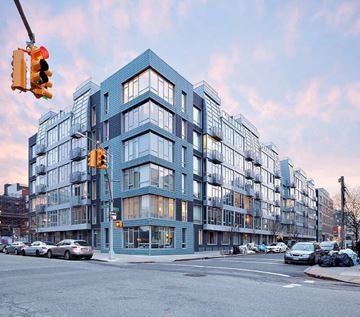 Brooklyn apartment for rent - A modern multi-story building with a blue tinted facade is situated on a street corner with cars parked in front.