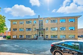Burlington apartment for rent - A black car is parked in front of a tan building with a glass door and windows.
