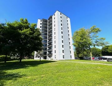 Mississauga apartment for rent - A tall white building with many windows is surrounded by trees and grass.