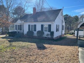 Fuquay-Varina house for rent - A white house with a black roof and a chimney is surrounded by trees and grass.