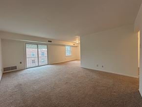 Fairless Hills apartment for rent - A large, empty room with carpeted flooring and a window with a view of a building outside.