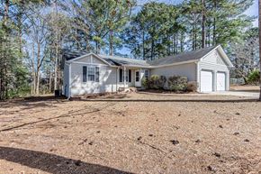 Midway Park house for rent - A house with a grey roof and white walls is surrounded by trees and has a brown dirt ground in front.