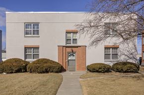Omaha apartment for rent - A building with a red door and windows surrounded by bushes and a tree.