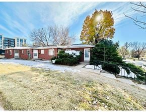 Denver apartment for rent - A small building with a red brick exterior is surrounded by a grassy area with a tree and a sidewalk.