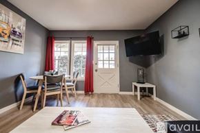 Los Angeles house for rent - A living room with a white door, a grey sofa, a wooden table and chairs, a flat screen TV, and a painting on the wall.