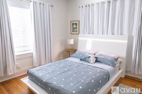 Miami apartment for rent - A bed with a blue comforter and white pillows is in a room with white walls and a window with white curtains.