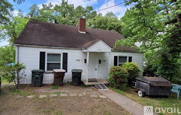 Greensboro apartment for rent - A white house with a brown roof and a chimney is surrounded by trees and has a trash can and a grill in front of it.