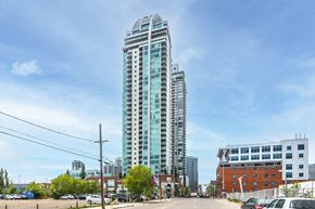 Calgary apartment for rent - A tall glass building stands in the middle of a parking lot.