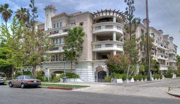 Sherman Oaks apartment for rent - A large, multi-story building with a balcony on the second floor is surrounded by greenery and a car is parked in front.
