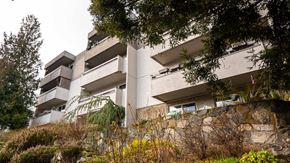 Victoria apartment for rent - A large white building with balconies and windows is surrounded by trees and shrubs.
