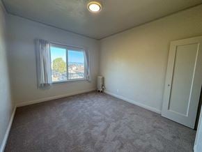 Alameda apartment for rent - A room with a carpeted floor, a window with a view of trees and buildings outside, a white door, and a ceiling light fixture.
