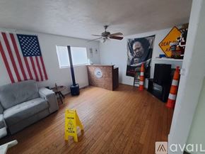 Kalamazoo house for rent - A living room with a grey couch, a wooden coffee table, a yellow caution sign on the floor, and a poster of a man's face on the wall.