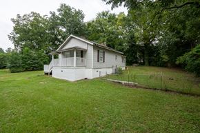 Fayetteville house for rent - A small house with a porch is surrounded by a green lawn and trees.