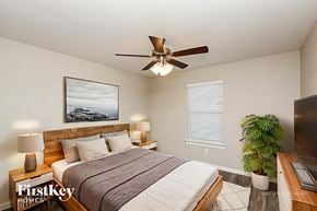 North Richland Hills house for rent - A bedroom with a bed, a ceiling fan, a nightstand with a lamp, a window with blinds, and a plant.