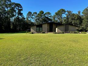Statesboro house for rent - A modern house with a flat roof and large windows is surrounded by a grassy field and trees.