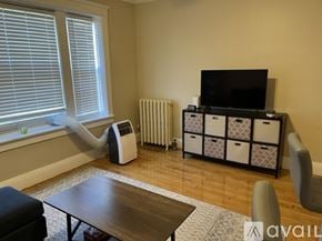 Boston apartment for rent - A living room with a television, a wood table, a sofa, a window with blinds, a radiator, and a cabinet with storage boxes.