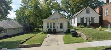 Omaha house for rent - A small white house with a black door and windows is surrounded by a green lawn.
