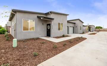 Visalia apartment for rent - A grey house with a black door and windows is surrounded by a gravel area and a concrete pathway.