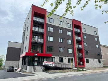 Fredericton apartment for rent - A multi-story building with a red and grey facade and a sign that says "Now Renting" and a phone number.