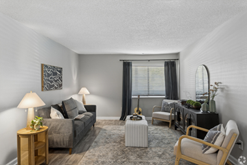 Nashville apartment for rent - A living room with a grey couch, a wooden side table with a lamp, a grey armchair, a white ottoman, a black cabinet with a plant on top, and a guitar leaning against the wall.