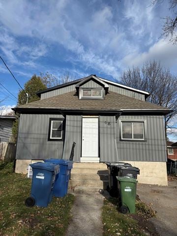 Guelph apartment for rent - A small house with a white door and two windows, one of which is covered with a screen, sits on a lawn with two trash bins in front.