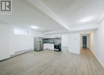 Toronto (South Parkdale) multi-family for rent - A spacious room with wooden flooring and a kitchen area with white cabinets and appliances.